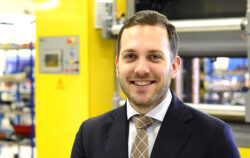 Smiling man in a suit and tie in an industrial factory setting with yellow machinery behind him.