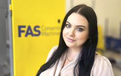 Portrait of a woman with long dark hair and blue eyes posing indoors in front of a bright yellow sign with blue text.