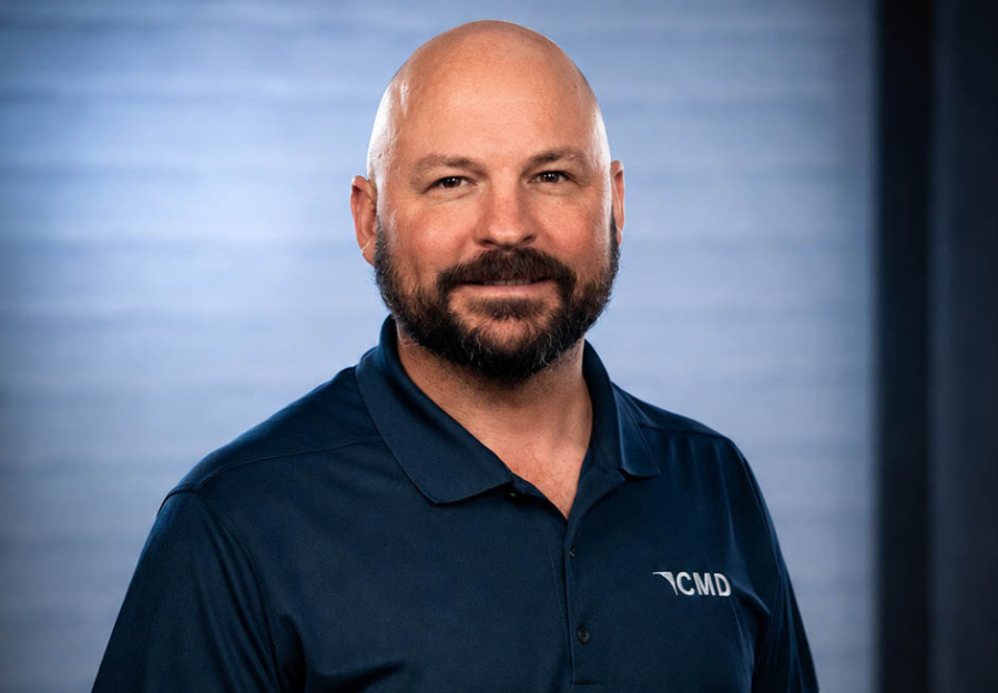 Head-and-shoulders portrait of a bald man with a beard, wearing a navy CMD polo, smiling against a blue background.